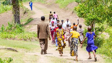 A group of adults and children walk along a dirt path in a rural, green landscape.