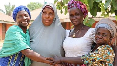 Four African women smiling and holding hands. They wear colorful headwraps and traditional clothing.