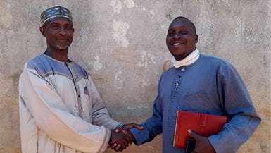 Two men shaking hands in front of a textured wall, one holding a red book.