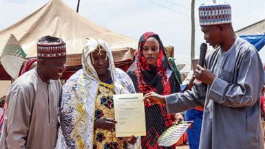 Group of people gathered for an event, one holds a document while another speaks into a mic.
