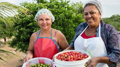 Duas mulheres do nordeste do Brasil sorrindo e segurando baldes de frutas frescas, com pequenas maçãs verdes e vermelhas