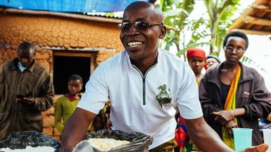 Um homem do Burundi vestindo uma camisa pólo branca e óculos de leitura sorrindo, estendendo as mãos e mostrando aos moradores do povoado como preparar mingau