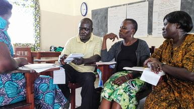 Tres mujeres y un hombre están sentados en un salón de clases en el norte de Uganda durante un taller de la Iglesia Asambleas de Dios Pentecostal, cada uno con un cuaderno a rayas