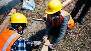 Dos trabajadores de la construcción, con cascos amarillos, cortan una madera por la mitad con serrucho para construir una vivienda antisísmica en Nepal