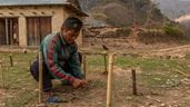 Um homem agachado no chão, em um povoado no Nepal, fazendo um gabião para impedir deslizamentos de terra