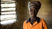 Une jeune femme d’une chorale en RDC porte une coiffe africaine et se tient devant la fenêtre d’une hutte en terre.