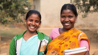 Dos mujeres que invierten en su comunidad local en Bangladesh por medio de un grupo de ahorros y préstamos, posan juntas sonriendo, una de ellas con libros en el brazo.