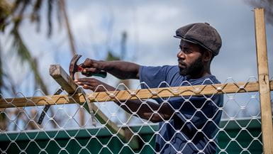Un hombre con gorra café y camiseta azul repara una construcción de su aldea de Siuiri, en Vanuatu