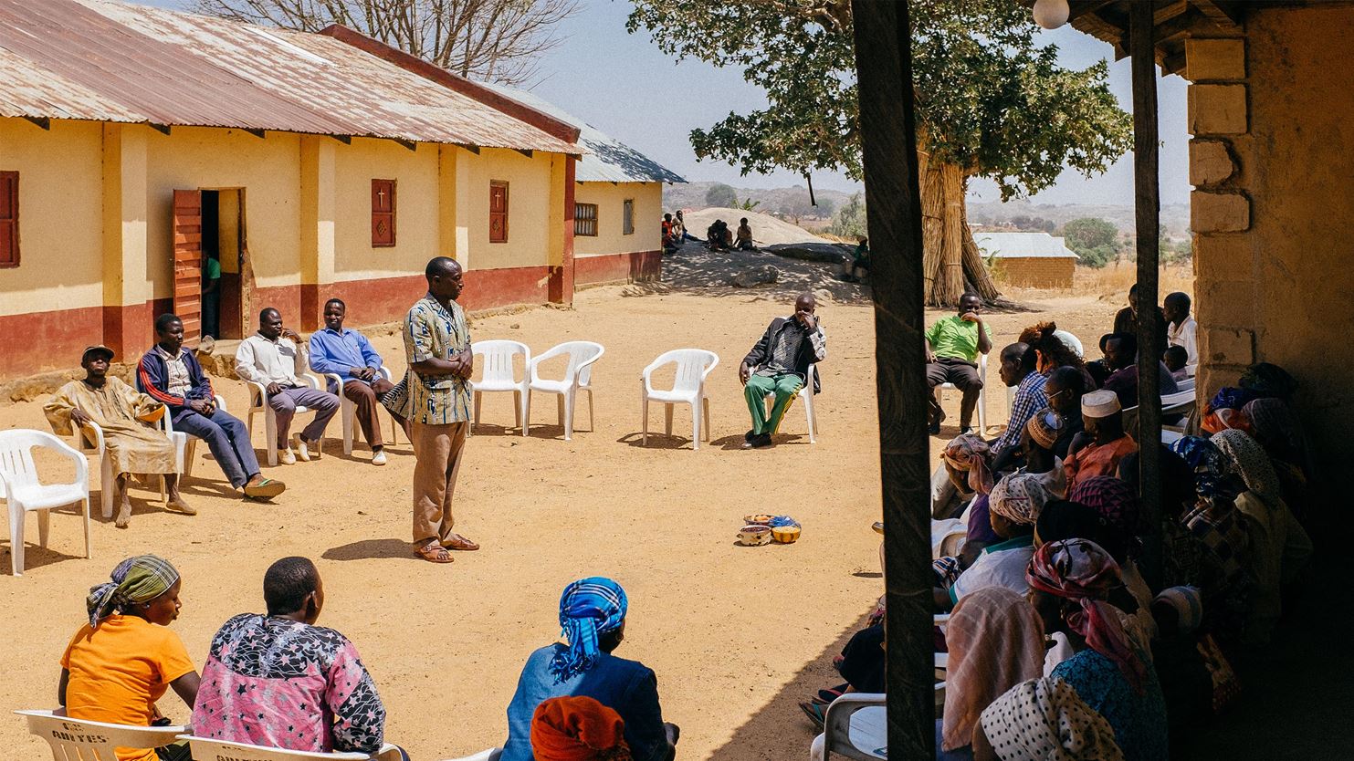 Um grupo de homens e mulheres na Nigéria sentados em cadeiras de plástico em círculo, ouvindo um facilitador no centro explicar sobre o Programa de Mobilização de Igrejas e Comunidades