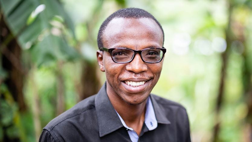 Photographie en gros plan d'un homme africain souriant portant des lunettes avec des palmiers bananiers en arrière-plan. Gilbert Irahari, agent de projet AEE-CCT. Kigali, Rwanda.