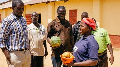Un groupe d’hommes et de femmes d’un petit village du Nigeria devant l’église dans une rue poussiéreuse, une femme tient un bol de pommes de terre et un homme tient une grande courge.